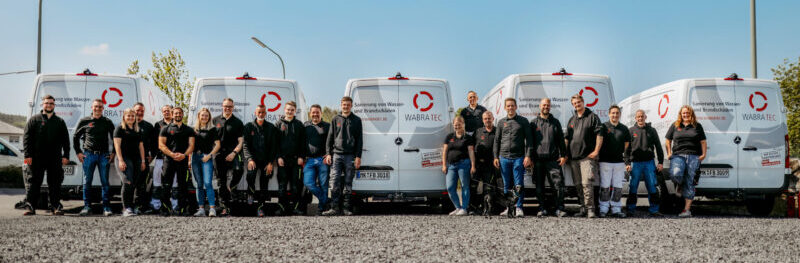 Das Team von WABRA TEC steht auf einem Schotterparkplatz vor den Firmenbullis und lächelt in die Kamera. Sie warten darauf die Fragen aus dem FAQ zu beantworten.