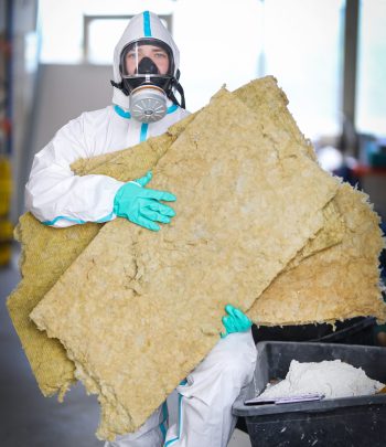 Ein Handwerker mit Atemmaske und Schutzanzug trägt während einer Schimmelpilzsanierung die kontaminierten Dämmung aus einem Objekt.