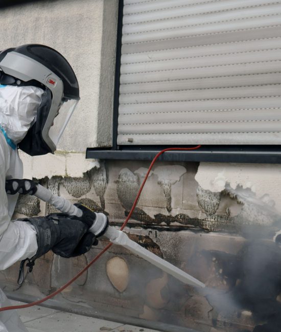 Ein Mitarbeiter der Firma WABRA TEC trägt Schutzkleidung und reinigt mit einem Trockenstrahlgerät für eine Brandschadensanierung eine Hauswand von Ruß.